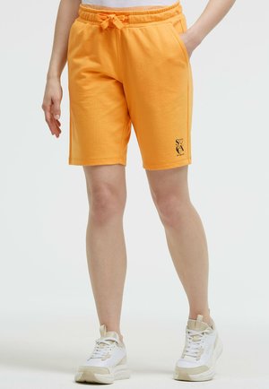 Person trägt orangefarbene Shorts mit Kordelzug und Seitentaschen sowie weiße Sneakers und steht vor einem einfarbigen Hintergrund.
