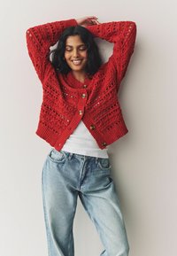 Cardigan en tricot rouge avec un motif ajouré, doté d'un large col et de gros boutons, porté sur un haut blanc et un jean bleu clair.