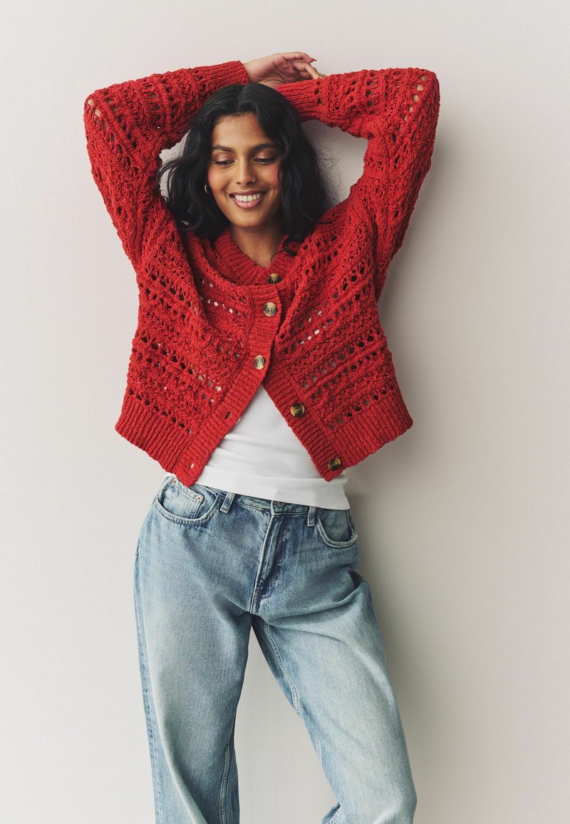 Cardigan en tricot rouge avec un motif ajouré, doté d'un large col et de gros boutons, porté sur un haut blanc et un jean bleu clair.
