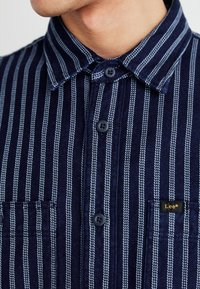 Närbild av en marinblå och vit randig skjorta med krage och en liten "Lee"-etikett på bröstfickan.