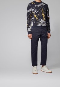 Navy sweatshirt with white and yellow palm leaf pattern, paired with dark trousers and white sneakers with a tan sole.