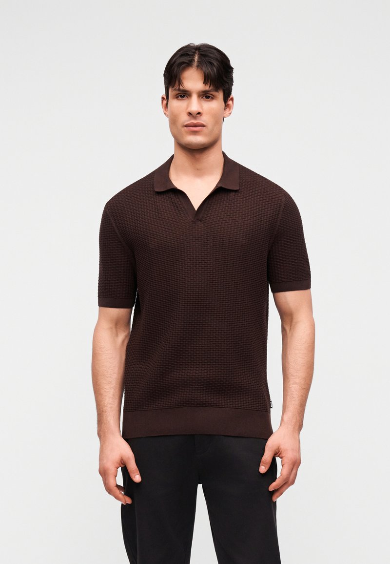 Άνδρας που φοράει σκούρο καφέ κοντομάνικο πόλο με υφή και μαύρο παντελόνι, στέκεται μπροστά σε απλό ανοιχτό γκρι φόντο.