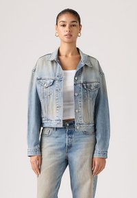 Γυναίκα που φοράει ανοιχτό μπλε ξεθωριασμένο τζιν μπουφάν και τζιν παντελόνι με λευκό κοντό τοπ, στέκεται μπροστά σε απλό ανοιχτό φόντο.