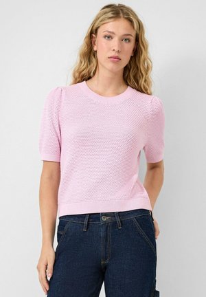 Młoda kobieta o falowanych blond włosach, ubrana w jasnoróżowy sweter z krótkim rękawem o teksturowanym wzorze oraz ciemnoniebieskie dżinsy, stojąca na neutralnym tle.