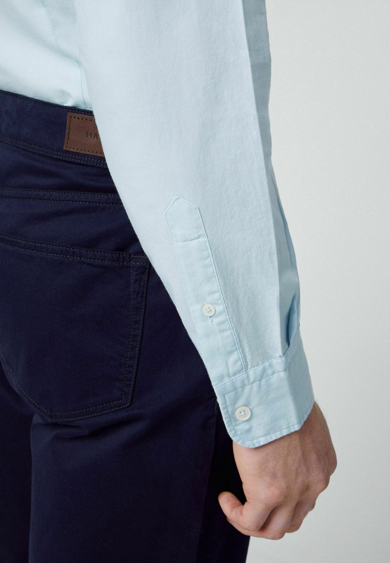 Hellblaues Langarmhemd mit Knopfmanschetten und spitzen Ärmeldetails, kombiniert mit dunkelblauen Hosen, die einfarbige Farben und ein schlichtes Design zeigen.