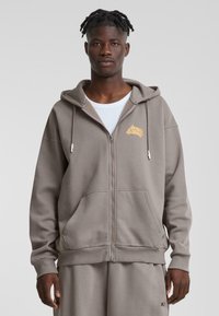 Sudadera gris con cremallera, con un gran bolsillo frontal, capucha con cordones y un logo naranja en el pecho. Tela suave y corte holgado.