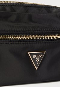 Must nylonist käekott kuldse luku ja kolmnurkse logoga, millel on tekst "GUESS" ja küsimärk. Siledapinnaline ja stiilse disainiga.