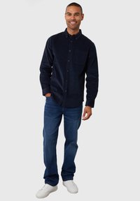 Mørkeblå skjorte med knapper i blød stof, med en brystlomme, parret med blå jeans og hvide sneakers, stående på en neutral baggrund.