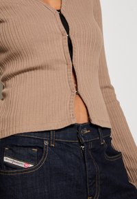 Ribbed beige cardigan with a V-neck, front zipper closure, and cropped design, paired with dark denim jeans featuring a side logo tag.