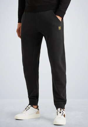 Homme portant un pantalon de jogging noir avec des poignets élastiques et des baskets blanches, se tenant avec une main dans sa poche contre un fond clair.