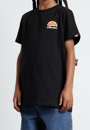 Osoba nosząca czarną koszulkę Ellesse z logo na piersi i rękawie, w połączeniu z niebieskimi dżinsami, na jasnoszarym tle.