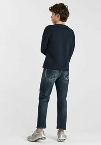 Marine sweatshirt, donkere denim jeans en zilveren sneakers. De outfit heeft een ontspannen pasvorm met zichtbare stiksels en textuurstof.