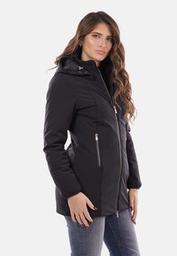 Giacca nera con zip e cappuccio, dotata di tasche frontali, dalla forma aderente e con una texture liscia. Dettagli in metallo argentato.