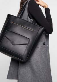 Sac fourre-tout en cuir synthétique noir avec une poche zippée à l'avant et un rabat plat repliable. Texture lisse, forme rectangulaire, couture minimaliste.