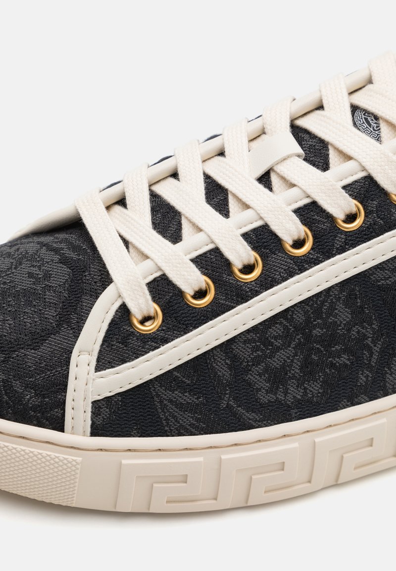 Sneaker in tessuto nero con texture floreale, lacci crema, occhielli dorati e suola in gomma bianca con un motivo geometrico.