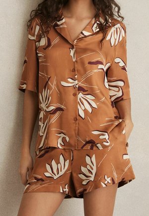 Femme portant une chemise à manches courtes boutonnée à motifs floraux bruns et un short assorti, debout devant un fond neutre.