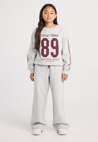 Girl standing wearing grey tracksuit with maroon stripes and "New York 89 Forever & Always" text, white sneakers, neutral background.