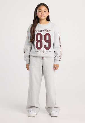 Fille debout portant un survêtement gris avec des rayures bordeaux et le texte "New York 89 Forever & Always", baskets blanches, fond neutre.