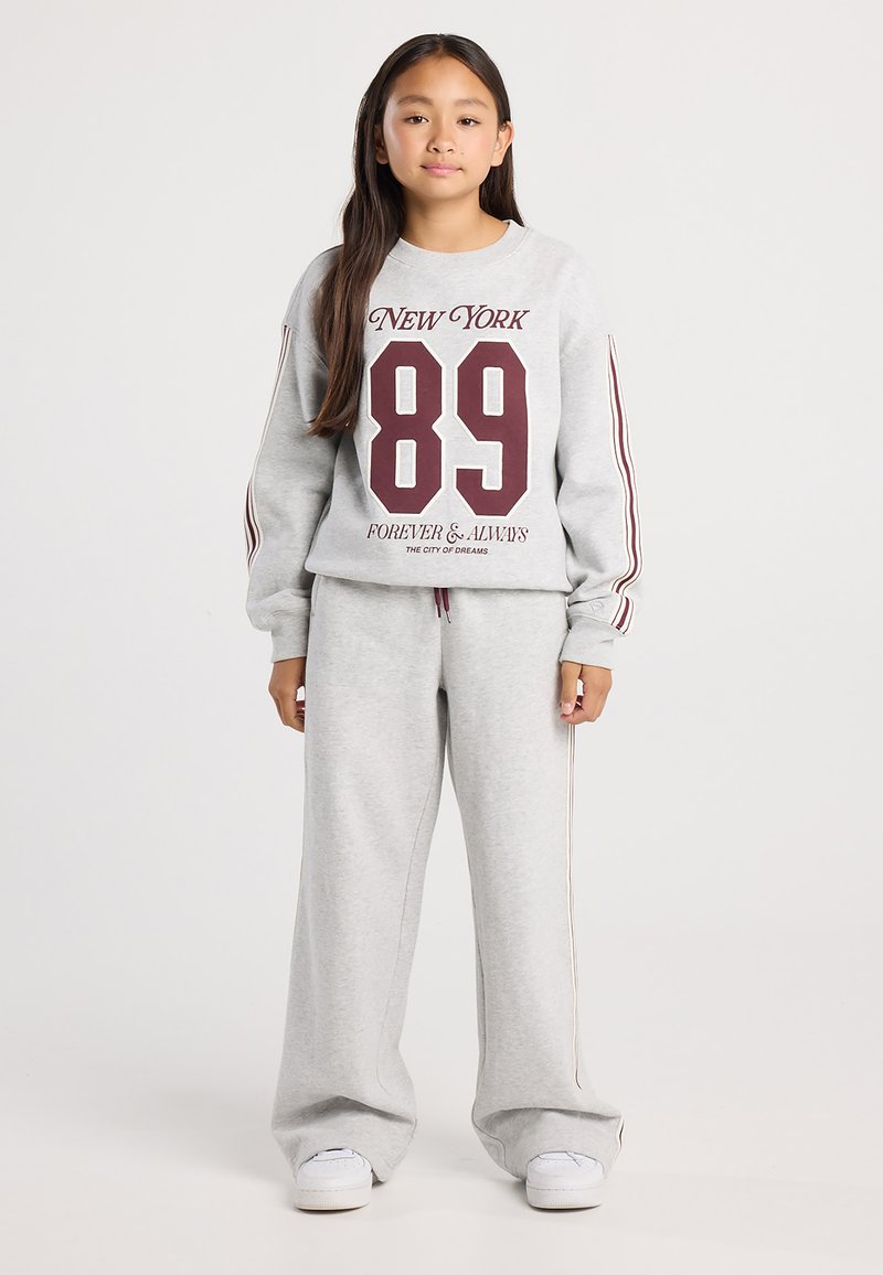 Girl standing wearing grey tracksuit with maroon stripes and "New York 89 Forever & Always" text, white sneakers, neutral background.