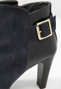 Gros plan d'une bottine noire en daim et cuir avec une sangle à boucle dorée et un talon haut empilé.