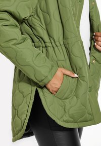 Giacca verde trapuntata con vita cintata, caratterizzata da una texture liscia e tasche laterali. Il design include un motivo arrotondato e chiusure a scatto.