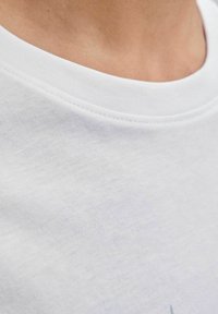 Close-up van de nek en bovenste borst van een persoon die een eenvoudige witte ronde hals T-shirt draagt met zichtbare stiksels.