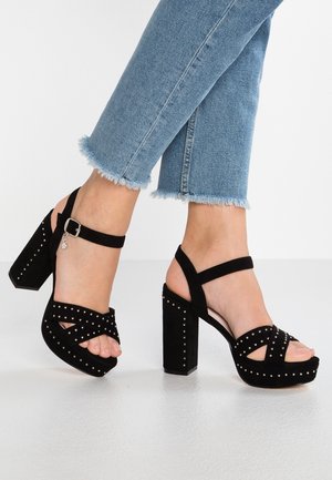 Füße in schwarzen, mit Nieten besetzten High-Heel-Sandalen kombiniert mit hellblauen, kurz geschnittenen Jeans mit ausgefransten Säumen auf einer hellen Oberfläche.