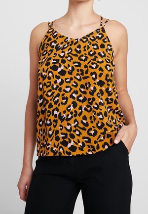 Vrouw draagt een mouwloos oranje shirt met zwarte en roze luipaardprint en zwarte broek, hand in zak.