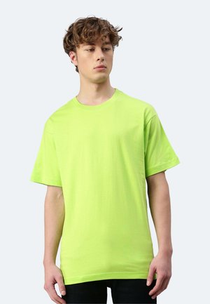 Jeune homme aux cheveux bouclés portant un t-shirt vert citron uni à manches courtes, debout devant un fond gris clair.