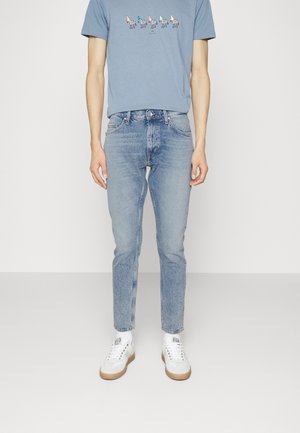 Man draagt lichtblauwe high-waist strakke jeans, een blauw T-shirt met kleurrijke dierenprints, witte sokken en witte sneakers met gumzolen.