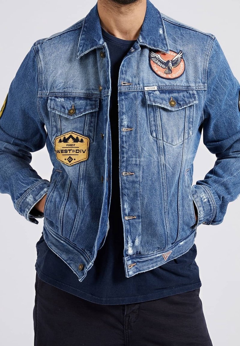 Muž v modré ošuntělé džínové bundě s lesními a orlími nášivkami, rukama v kapsách, přes tmavě modrou košili a černé kalhoty.