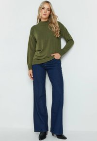 Maglione a maglia verde oliva con colletto alto, abbinato a jeans a coscia larga blu navy e stivali neri alla caviglia. Vestibilità casual con maniche lunghe.