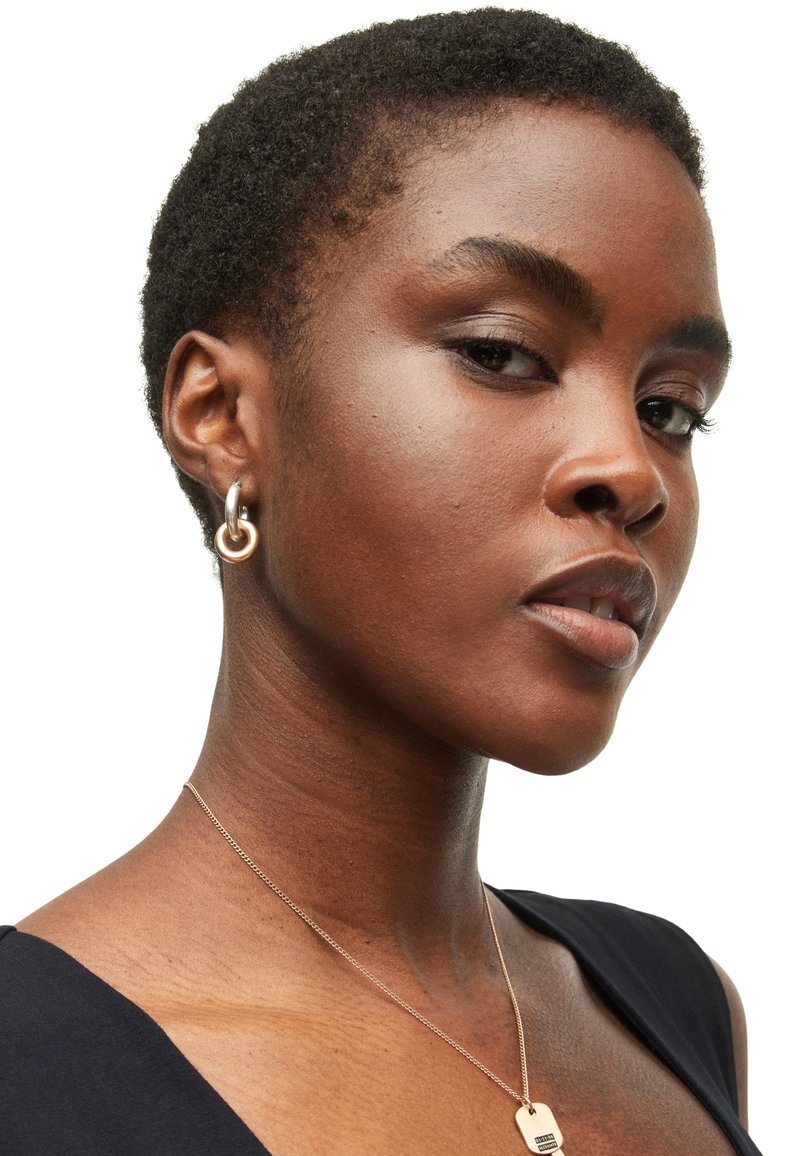Pendientes de aro de oro y plata con un diseño circular. Pelo corto y texturizado, lleva un collar de oro sencillo. Prenda negra. Fondo neutro.