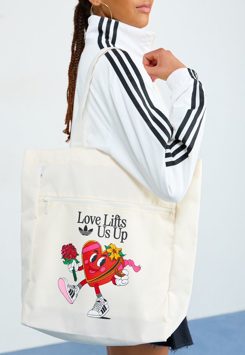 Canvas tote bag in cream, featuring a cartoon heart character, flowers, and the text "Love Lifts Us Up." Includes a zippered pocket.