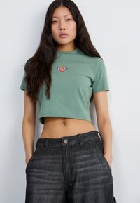 Green cropped t-shirt in soft fabric with short sleeves, featuring a multicolored logo on the chest. Paired with dark gray denim pants.