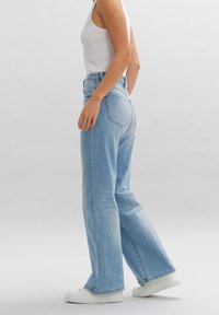 Jean taille haute bleu clair avec une coupe évasée. Confectionné en denim, il comporte des poches arrière et un effet délavé. Porté avec des baskets blanches.