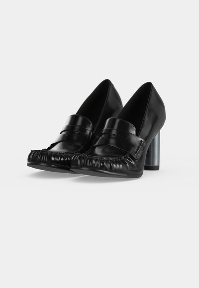 Paire de chaussures à talons hauts en cuir noir avec bouts arrondis et détails froncés à l'avant, sur fond blanc.
