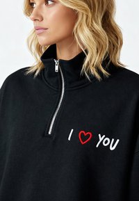 Sweat-shirt noir avec un col montant et une fermeture éclair à l'avant. Présente la broderie "I ♥ YOU" en rouge et blanc. Tissu doux avec une coupe décontractée.