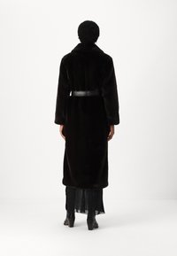 Largo abrigo de piel sintética negra con cintura ceñida por un cinturón, cuello muesca y mangas largas, combinado con una falda negra en capas y tacones.