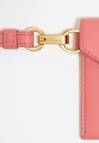 Różowa skórzana torba crossbody z paskiem, wyposażona w złote akcenty, zapięcie oraz prostokątną etykietę z wytłoczonym logo.
