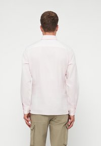 Camisa de manga larga color rosa claro, hecha de un tejido suave, con cuello clásico y puños abotonados, vista desde la parte de atrás. Combinada con pantalones cargo beige.