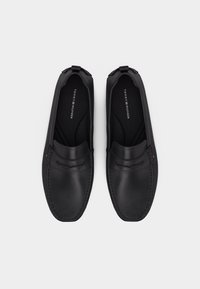 Ζευγάρι μαύρα δερμάτινα μοκασίνια χωρίς κορδόνια με ραφές και λογότυπο Tommy Hilfiger στο εσωτερικό, σε ανοιχτό γκρι φόντο.