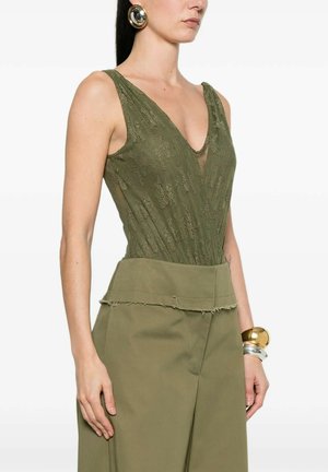 Donna che indossa un top senza maniche in pizzo verde oliva con scollo a V profondo, pantaloni a gamba larga e vita alta color oliva, grandi orecchini rotondi dorati e braccialetti robusti.