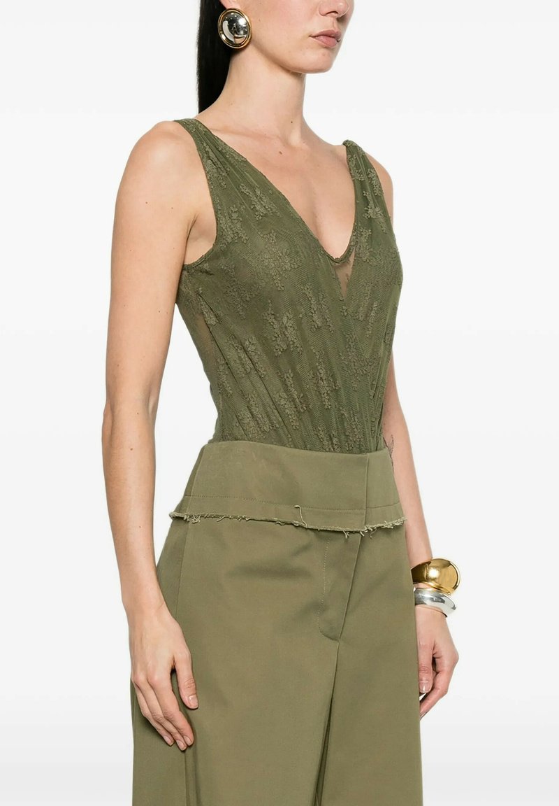 Donna che indossa un top senza maniche in pizzo verde oliva con scollo a V profondo, pantaloni a gamba larga e vita alta color oliva, grandi orecchini rotondi dorati e braccialetti robusti.