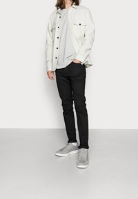 Ljusgrå skjorta över en grå t-shirt, matchad med svarta jeans och grå sneakers med vita sulor, stående på ett ljust golv.