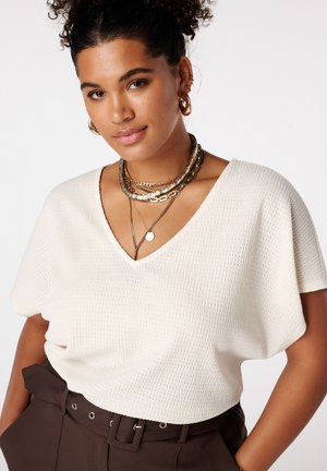 Vrouw met krullend haar die een crèmekleurige, gestructureerde V-hals top draagt, gelaagde gouden kettingen en donkere, met riem afgesloten broek met handen in de zakken.