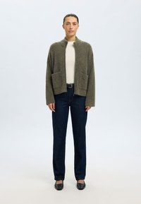 Volneno jopico olivno zelene barve z visokim ovratnikom, dvema sprednjima žepoma in sproščenim krojem. Nošena preko svetlejšega topa in temnih kavbojk.