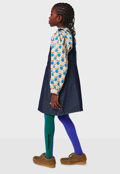 Robe salopette en denim portée sur un chemisier à motifs avec des cercles bleus et orange. Leggings colorés : verts et violets, avec des chaussures marron.