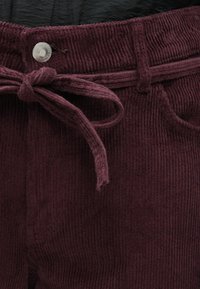 Artimas tamsiai violetinių kordūrinių kelnių vaizdas, su audiniu pasirinktu diržu, surištu į لویą, ir metaliniu mygtuku ant juosmens.