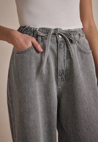 Pantalons en denim gris avec une taille élastique, un cordon de serrage, et des poches latérales. Le tissu a une texture lisse et une apparence délavée.